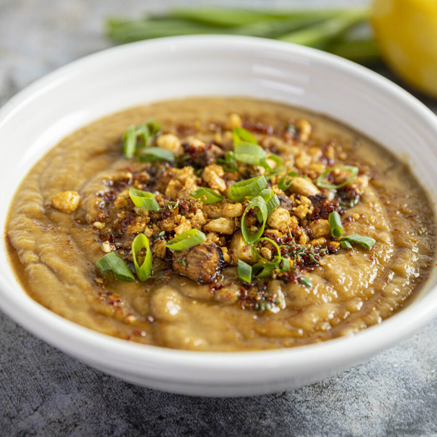 A bowl of creamy plant-based parsnip and fennel soup topped with spring onions, spices, and nuts, with lemon and spring onions in the background.