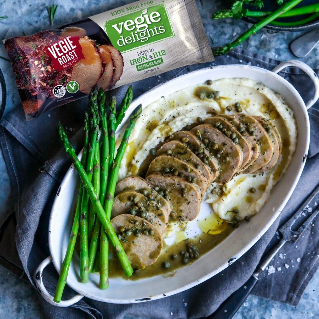 A plant-based roast served up in a baking dish, decorated with asparagus. A Vegie Delights packet sits just in shot.