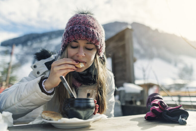 A woman wearing snow attire eats soup with crusty bread at a seat outside surrounded by snowy mountains.