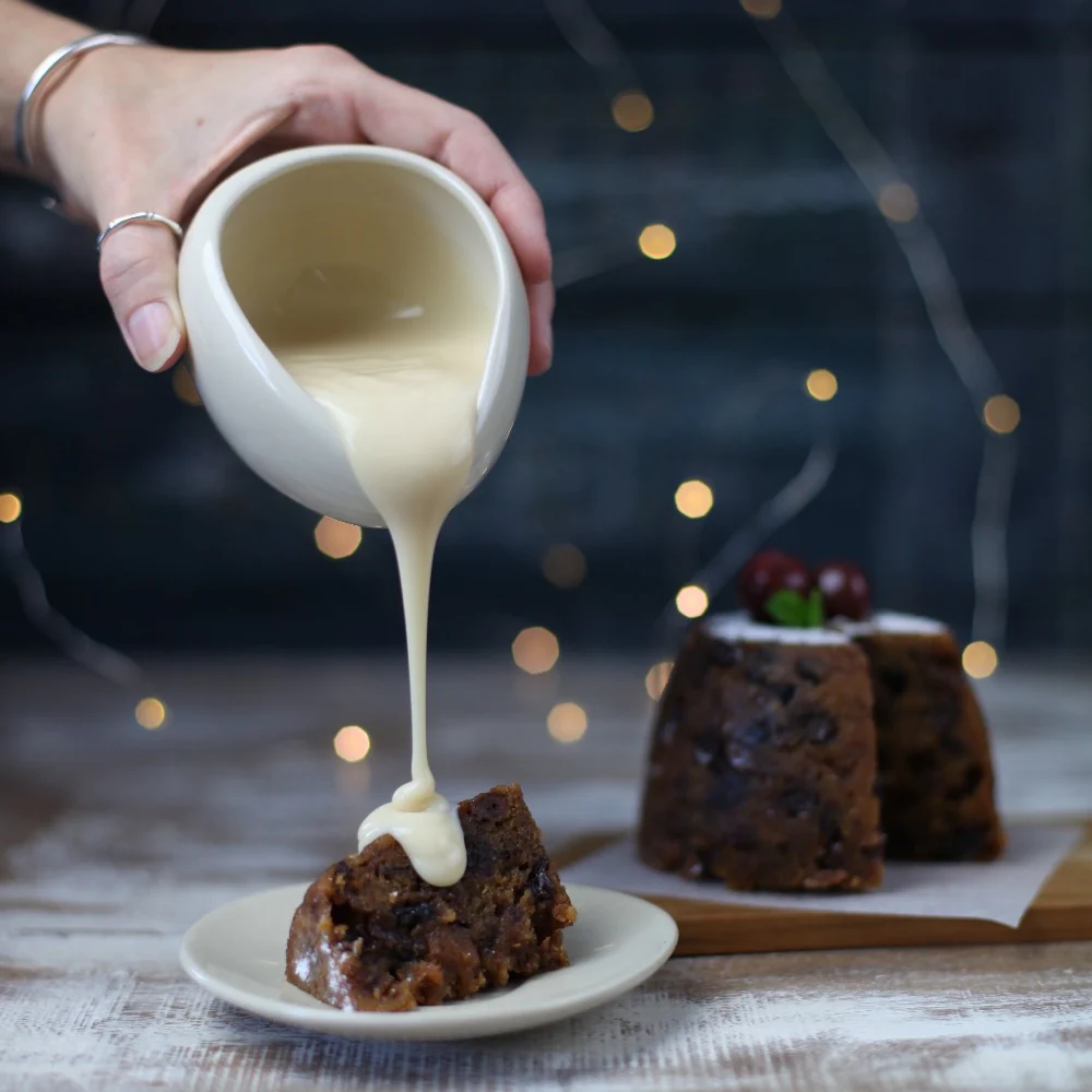 A slice of pudding on a plate with a hand pouring custard over the top.
